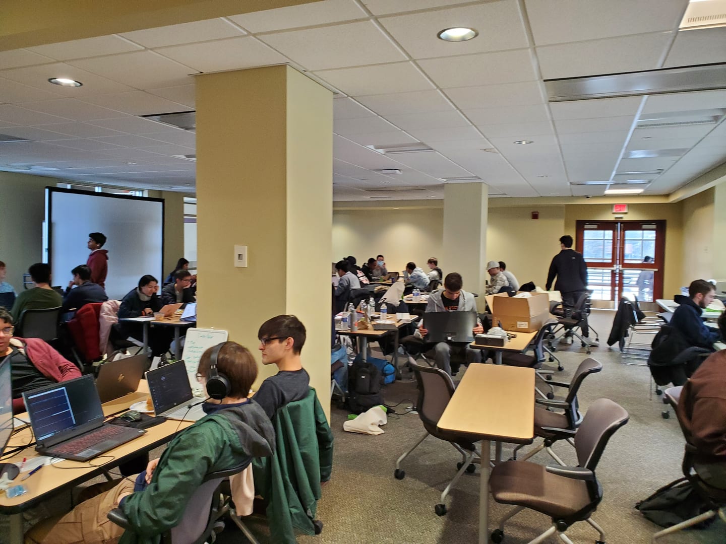 Students working on laptops at tables spread through a large study room, one wearing headphones in the foreground.