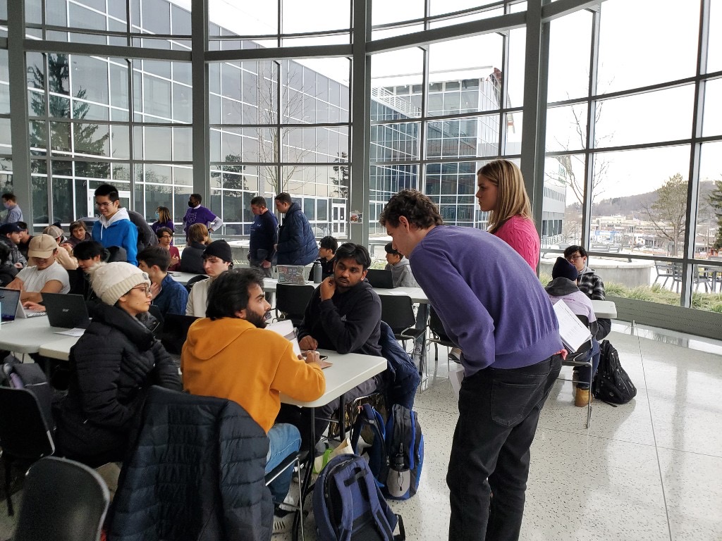 Students collaborating at workshop tables with laptops in a bright room with floor-to-ceiling windows at a HackBU event.