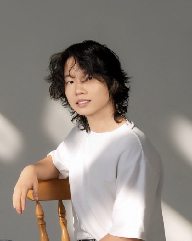Portrait of Raymond Chen, HackBU organizer, seated on a wooden chair in a white T-shirt against a sunlit gray wall.