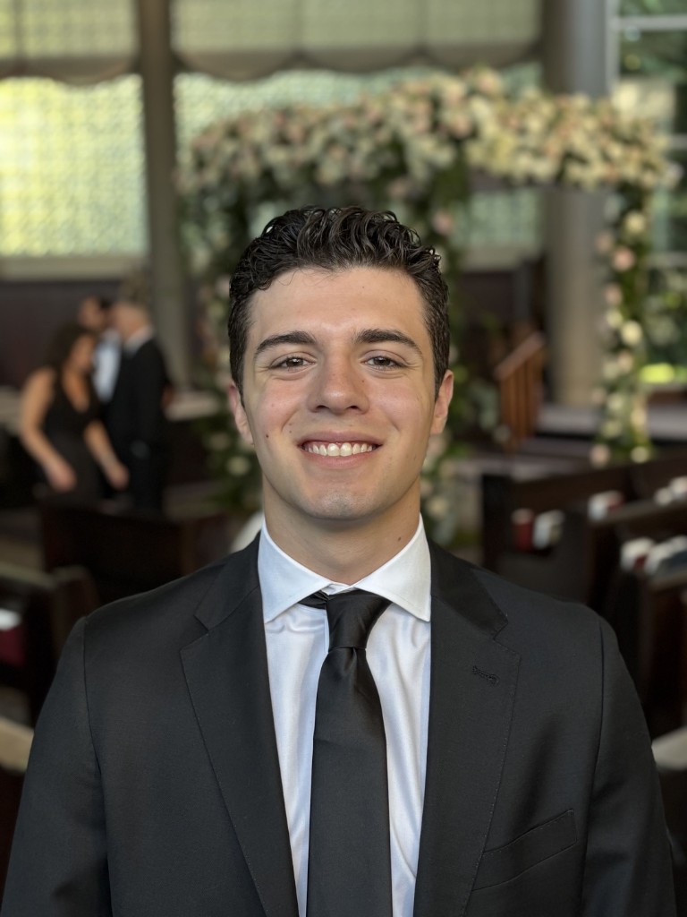 Portrait of Thomas Mandel, HackBU organizer, smiling in a dark suit and tie in front of a floral arch.