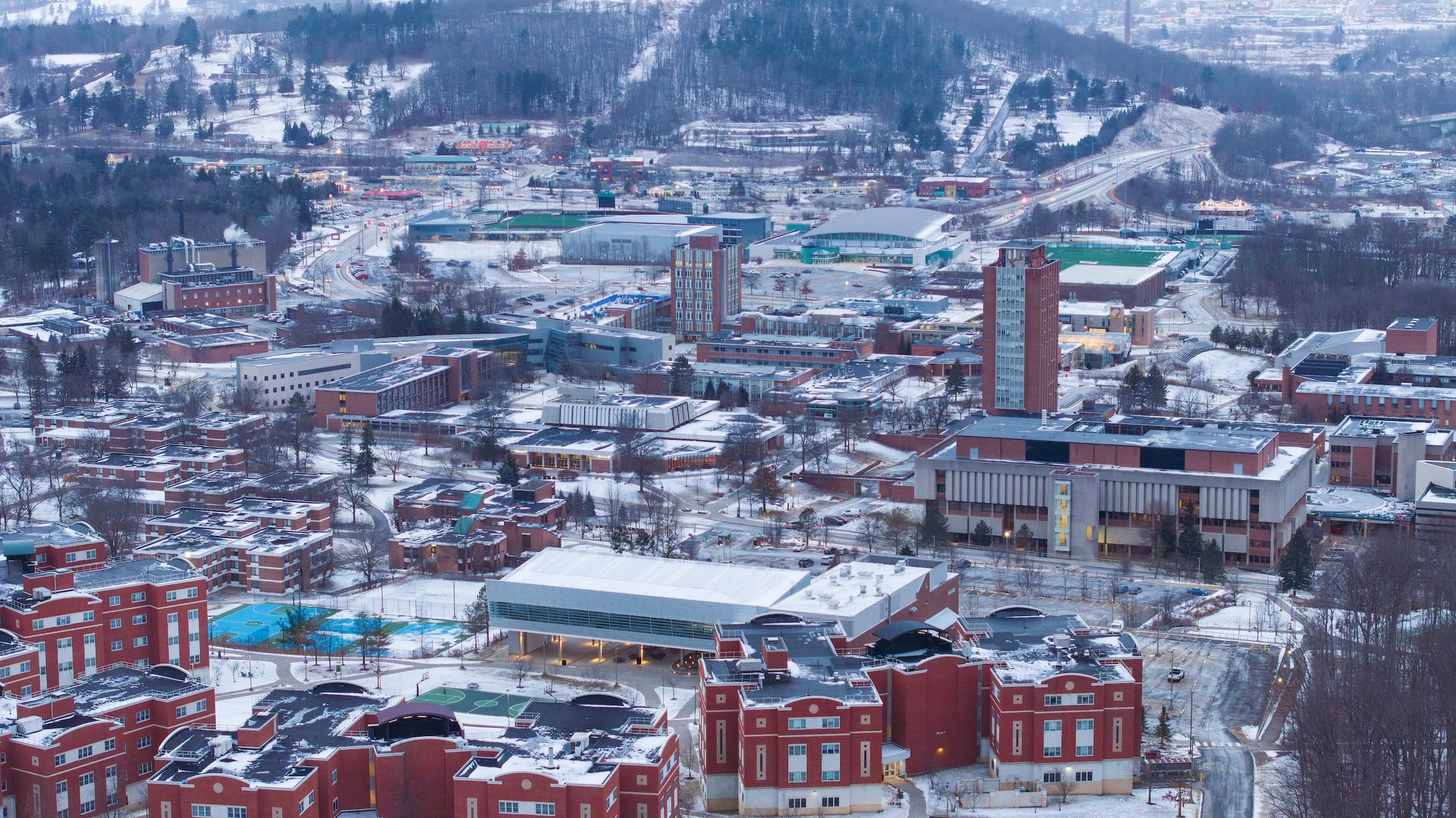 Aerial photograph of the Binghamton University campus in winter at dusk: the brick Library Tower rising on the right, snow-dusted academic buildings and red-brick residence halls below, athletic fields and wooded hills beyond, and lights coming on across campus.