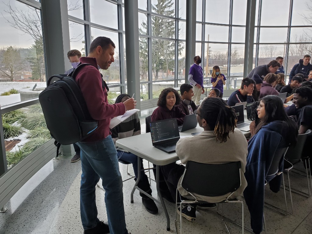 Students around a workshop table with laptops, talking with a mentor, winter campus visible through the windows.
